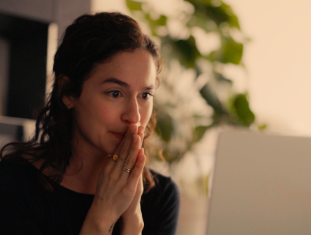 Close-up of a woman with long, curly hair, wearing various rings, pressing her hands together in front of her face while staring at the screen of a laptop
