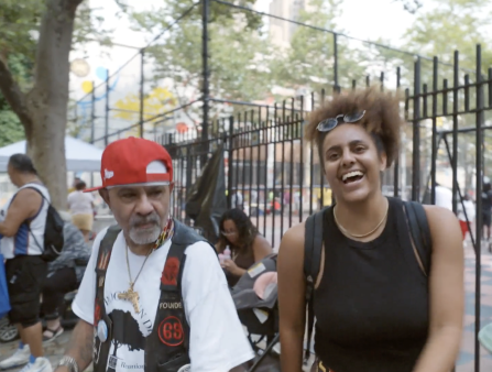 An older man in a white T-shirt, black vest, red hat, and golden gun-shaped necklace stands next to a laughing woman in a black tank top and backpack. The two are in front of a large New York City park, with trees, a wrought-iron fence, and a basketball court in the background.