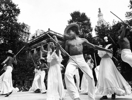 Young shirtless men in brimmed hats, flared white pants, and ballet flats hold long poles and dance alongside young women in white dresses on a stage in a park.