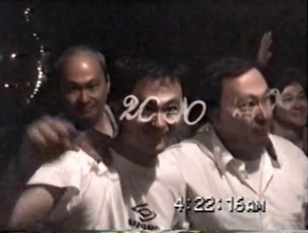 Low-resolution medium close of four men. The two in the front wear white T-shirts and glasses shaped like the number 2000. A timestamp at the bottom right corner of the screen reads 4:22:16AM