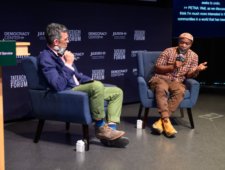 Two men sit in blue armchairs and converse on a stage, both holding microphones which read "ida international documentary association." Behind the men is a black background with the words "TATEUCHI DEMOCRACY FORUM", "janm", and "DEMOCRACY CENTER", and a screen displaying the text of their words. On the left is a man in a blue suit jacket and green pants (Dominic Asmall Willsdon), and on the right is a man in a matching brown, white, and orange patterned shirt and hat (Petna Katondolo)