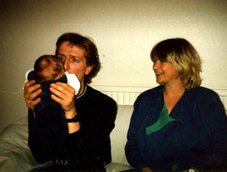 A man holds up a baby in front of his face and makes a silly expression. A woman sits next to him and smiles. Both wear dark clothing and sit on a sofa
