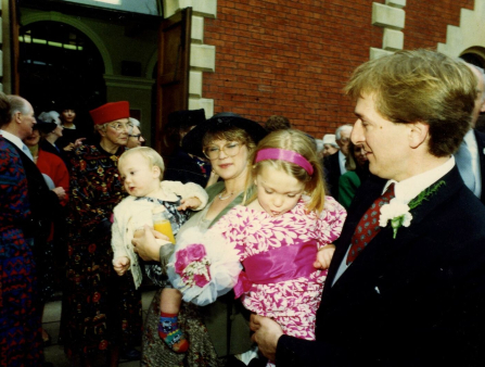 A man in a black suit and red tie with a white flower on his lapel holds a young girl in a pink floral dress in his arms and smiles at her. Behind the two a crowd mills about in front of a red brick building, including a woman in a green dress and a black hat holding a young boy in a white jacket. All of the crowd wear bold patterns and vibrant colors, and many wear hats