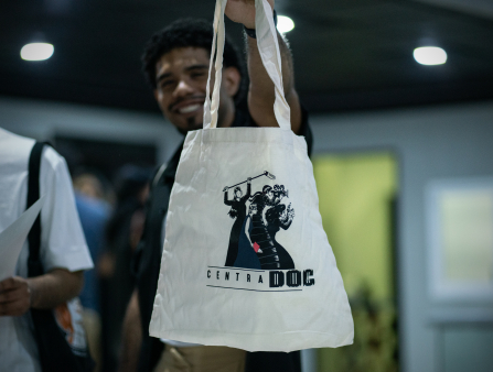 A smiling man holds a tote bag up to the camera, which is printed with a graphic of women filmmakers and text reading "CENTRADOC"