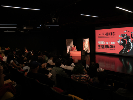 A sleek, black theater filled with a crowd watches a woman on stage behind a red podium speak. Two banners stand behind her. A screen on the stage displays the words "CENTRADOC PIONERAS EN EL CINE PROGRAMACIÓN" and a graphic of women filmmakers over a red background