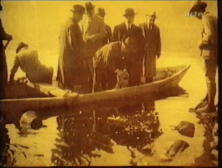 Yellow image of six men in coats and hats standing on a canoe and peering into the water beneath them. Another similarly dressed man sits on the boat, and another man stands on the shore and peers into the water himself