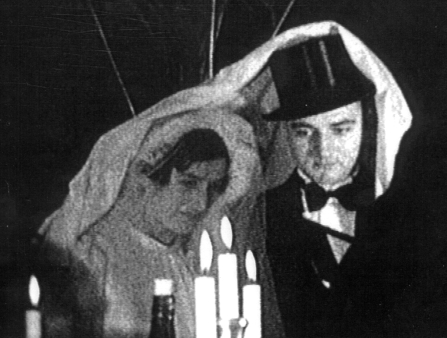 Low-resolution black and white image of a bride and groom looking somber with a white cloth over their heads. Three candles sit in front of the pair. The groom wears a tall hat