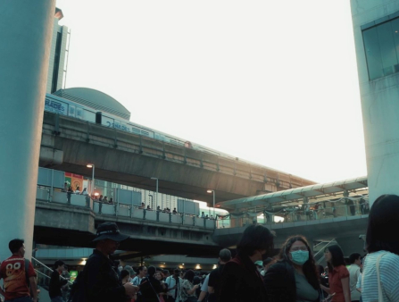 Shot of a train station from below with a crowd of people milling about on the ground. Tall buildings frame the image on either side. The sky is white