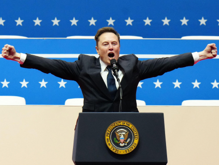 Musk speaks into a microphone on a podium with the Seal of the President of the United States. He wears a black suit and a blue tie, his arms are oustretched to either side and his hands form fists. The background is two rows of white stars over blue