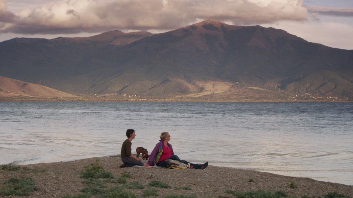 Two women with short hair, and a dog, sit in front of a lake and mountains