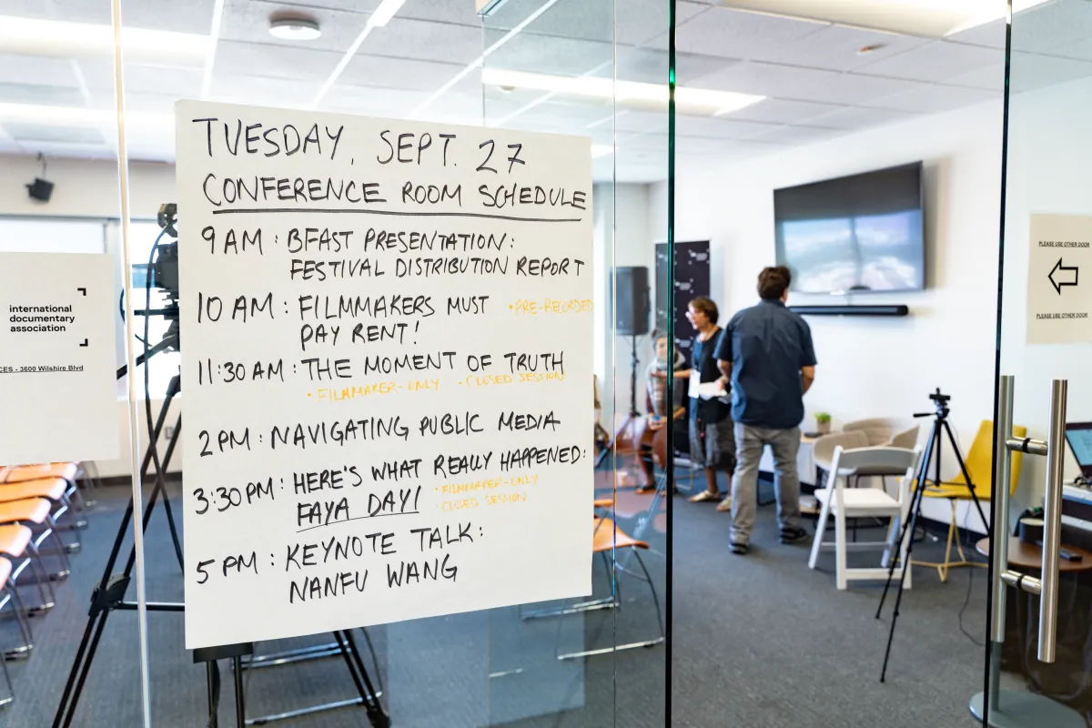 Conference room with yellow chairs, camera equipment and two people in the background wearing dark blue tops and gray bottoms. Sign on glass door in foreground with Conference Room schedule. 