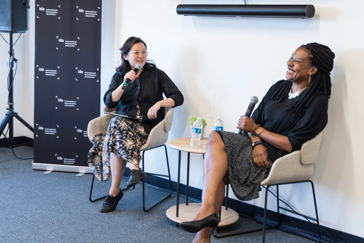 Two people sitting in chairs. Person on left is holding a microphone up to their face, wearing a floral skirt, black sweater, and black shoes. Person on right is holding a microphone down with a big smile, wearing a black and white polka dot skirt with a black sweater and black heels.