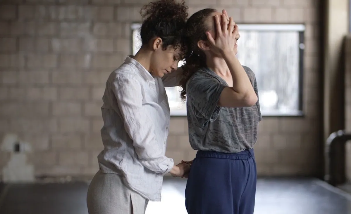 This photograph shows dancer Mor Mendel closely behind dancer Hadar Ahuvia, the two facing towards the right in an industrial looking dance studio with windows. They hold opposite hands behind Hadar's back, while the other pair of opposite hand's covers Hadar's eyes. Hadar's head leans back into Mor's forehead— they are contemplative. 