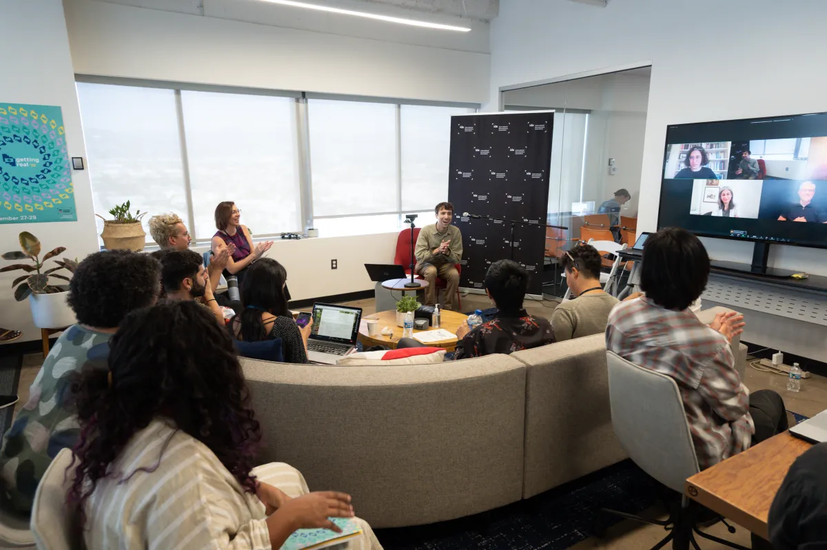 Photograph of a room filled with multiple people seated and clapping in applause. The focus is on Chris Boeckmann, Getting Real '22 Programmer, who is wearing brown pants and a light olive green long-sleeved button-up, sitting in front of a black backdrop.