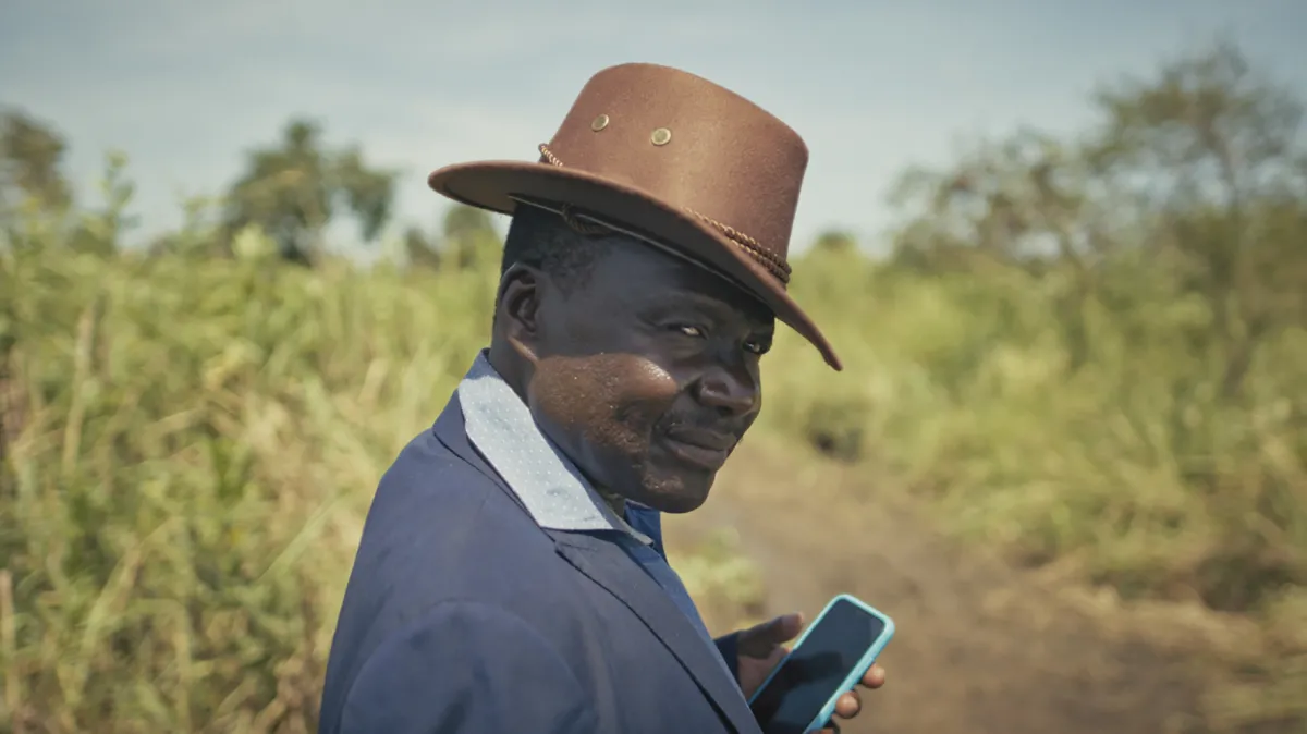 Photo courtesy of CPH:DOX. Still from 'Theatre of Violence.' A person with dark skin, wearing a brown rimmed hat and a blue coat, holding a cellphone, stands in a green field with a dirt path.