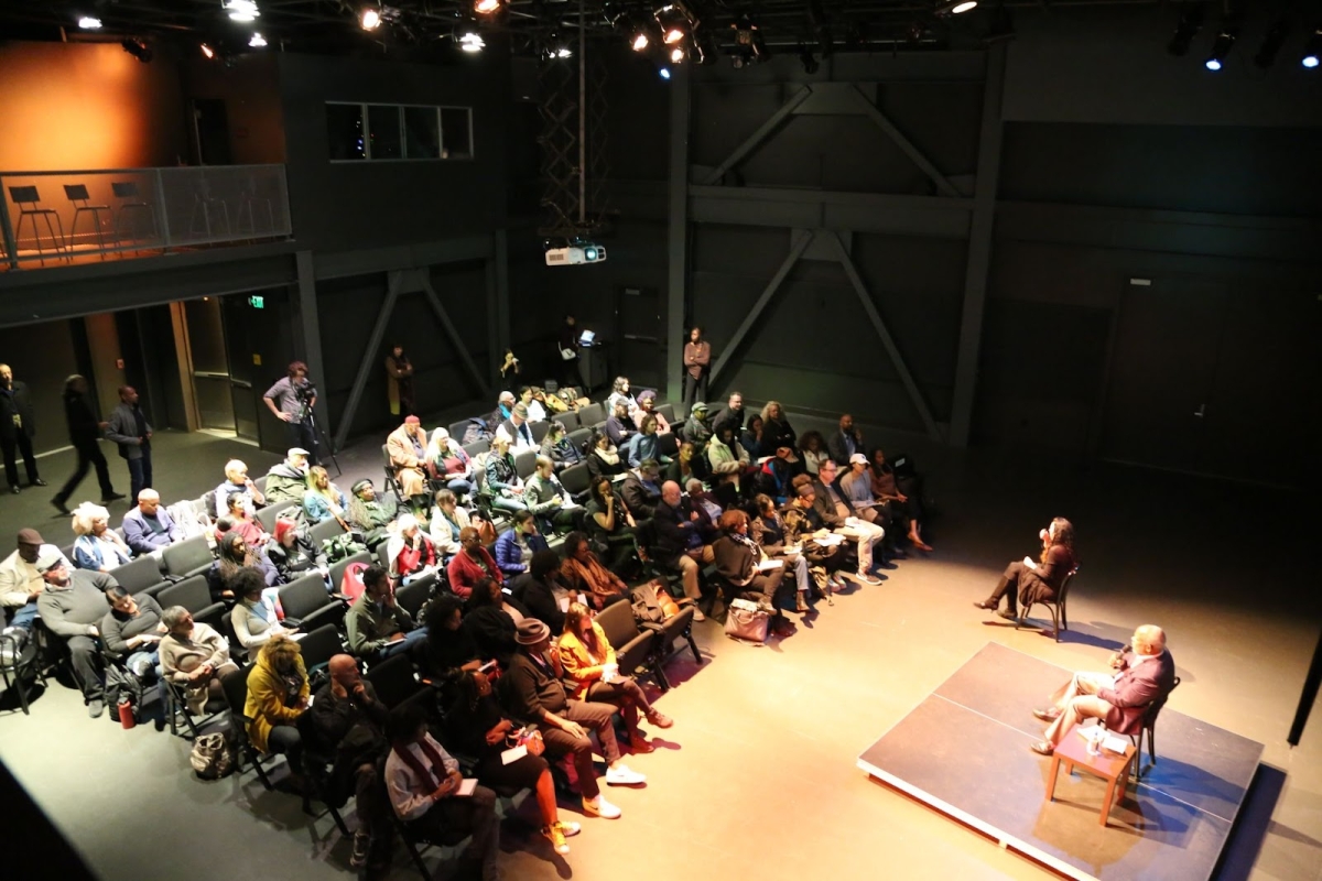 The wide angle image shows an group of more than 100 people are sitting facing Stanley Nelson who has a microphone in hand is presenting with light spotlighting him.