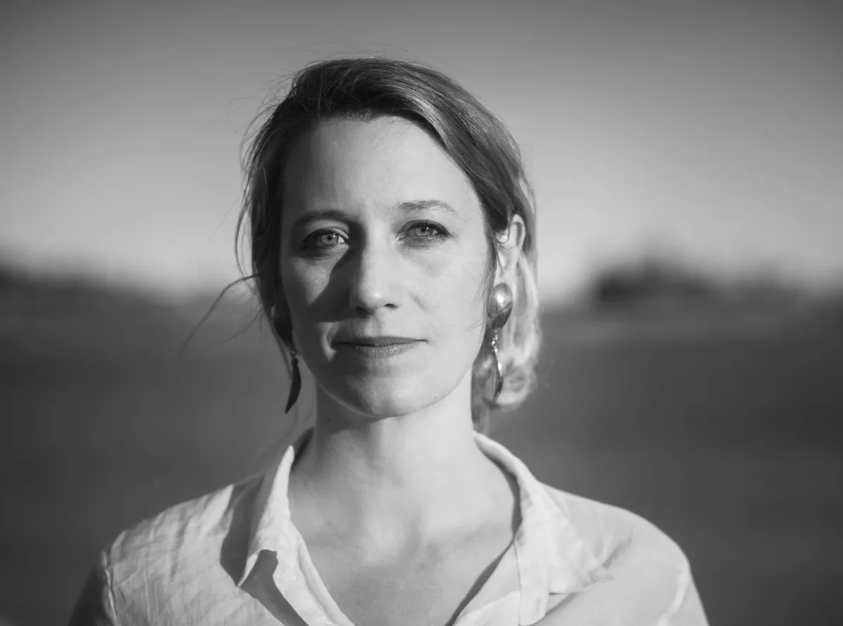 headshot of a white woman in her forties with hair loosely tied back, a white button down shirt, and metallic earrings standing in front of a body of water.