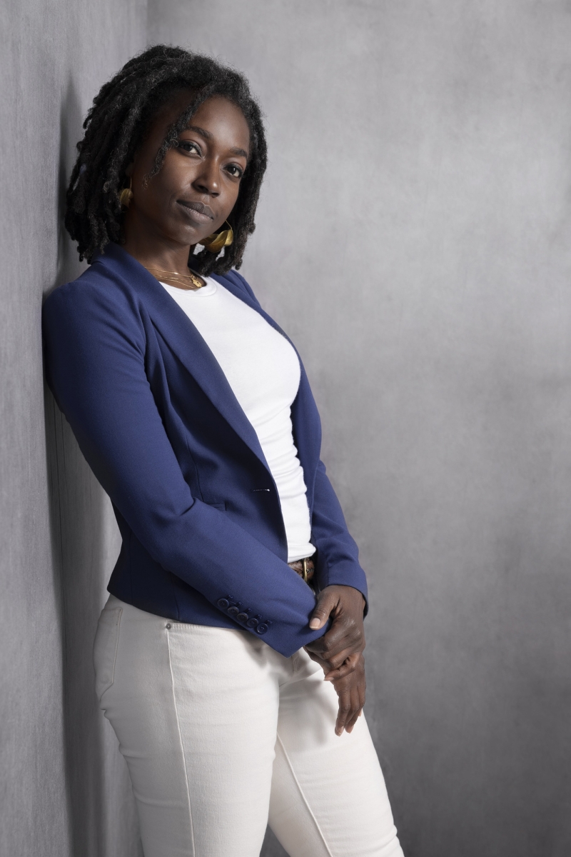 Black woman with shoulder length braids, standing against a gray wall. She is wearing a dark blue blazer with a white shirt and white pants. 