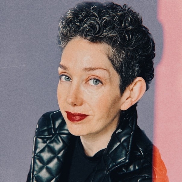 A woman with white skin smiles at the camera. She has short, curly salt-and-pepper hair, blue eyes, red lipstick, and is wearing a quilted black leather blazer. 