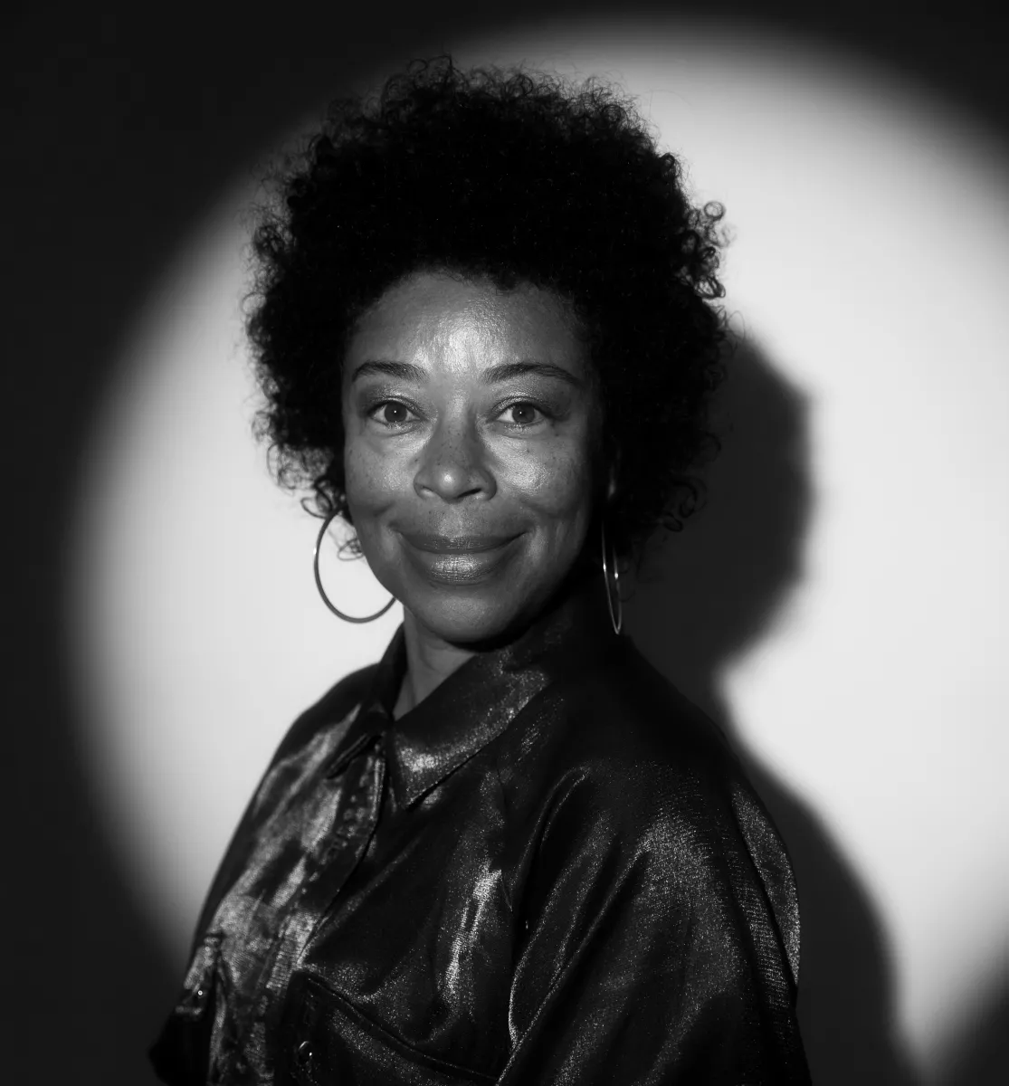 Black and white photo of a Black woman lit with spotlight with dark curly hair, wearing hoop earrings.