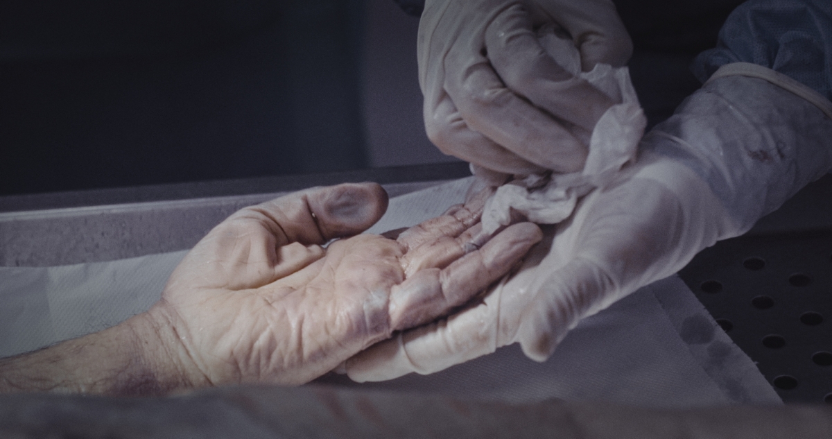 Still image from 'Pure Unknown,' depicting a pair of gloved hands cradling and gently wiping the hand of someone who is deceased.