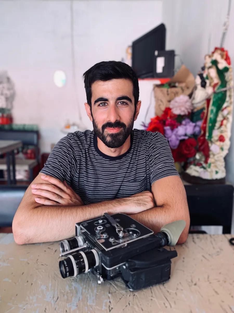 A person with fair skin and thick dark short hair, eyebrows and a short beard sit at a table looking gently at the camera with a smile. They cross their arms, in front of them is a 16mm camera.