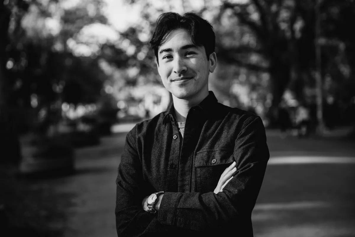 Black and white photo of a young man with fair skin and black hair and eyebrows. He is standing in an environment with trees behind him. He crosses his arms gleefully. He wears a button up collard shirt. 