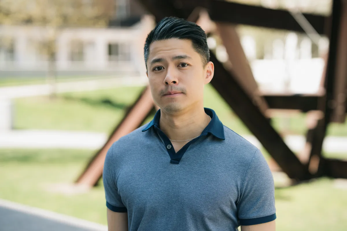 Headshot of adult of East Asian descent with medium-light skin; short hair; dark blue polo shirt; in front of grassy field