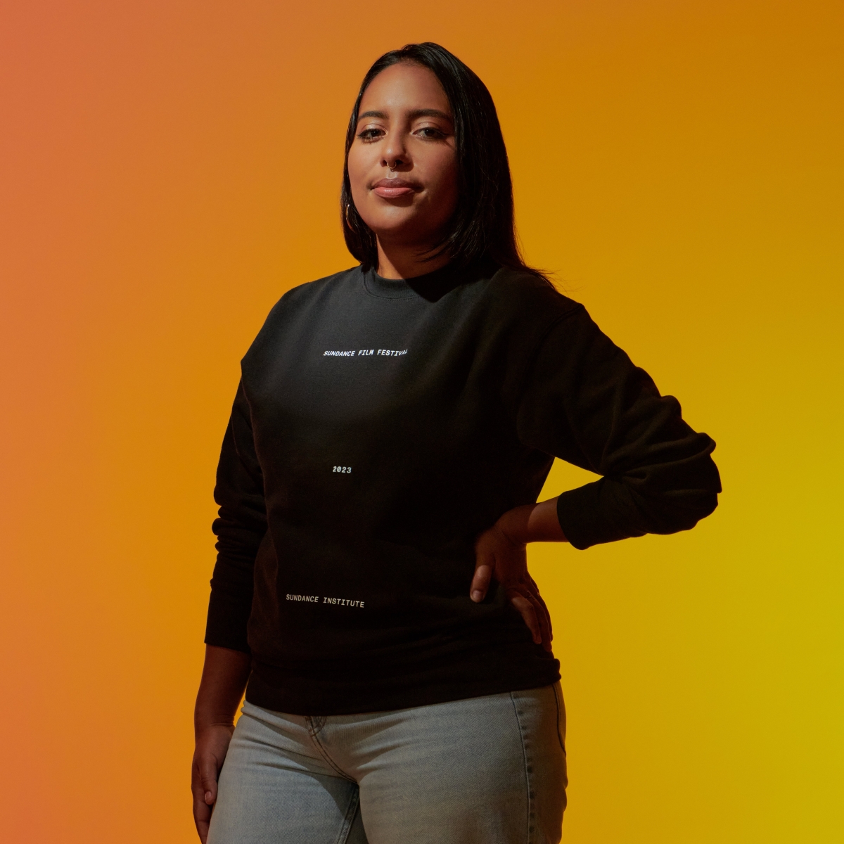 Headshot of Amber Espinosa-Jones, who is wearing a 2023 Sundance sweatshirt while standing in front of a yellow-orange background.