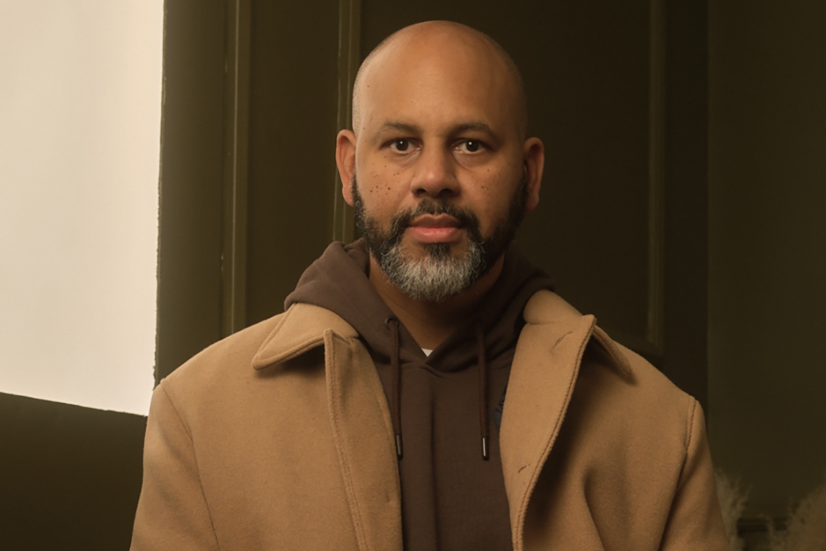 Headshot of an adult male with medium-dark skin tone, bald-headed, with a short beard, wearing a tan overcoat over a brown hoodie. 