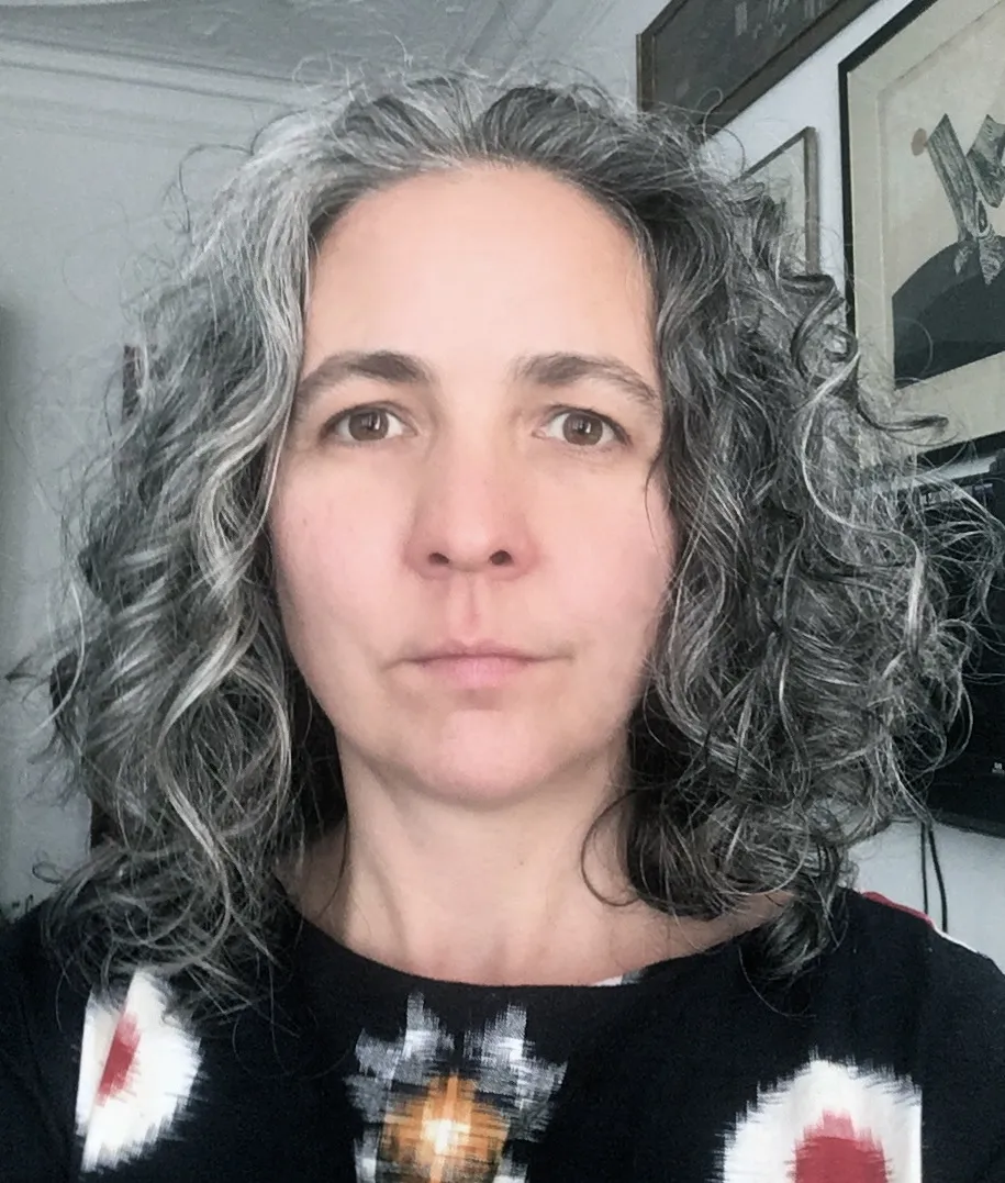 Headshot of a woman with grey/ brown curly hair and light skin, wearing a dark blue top with a brightly colored pattern.