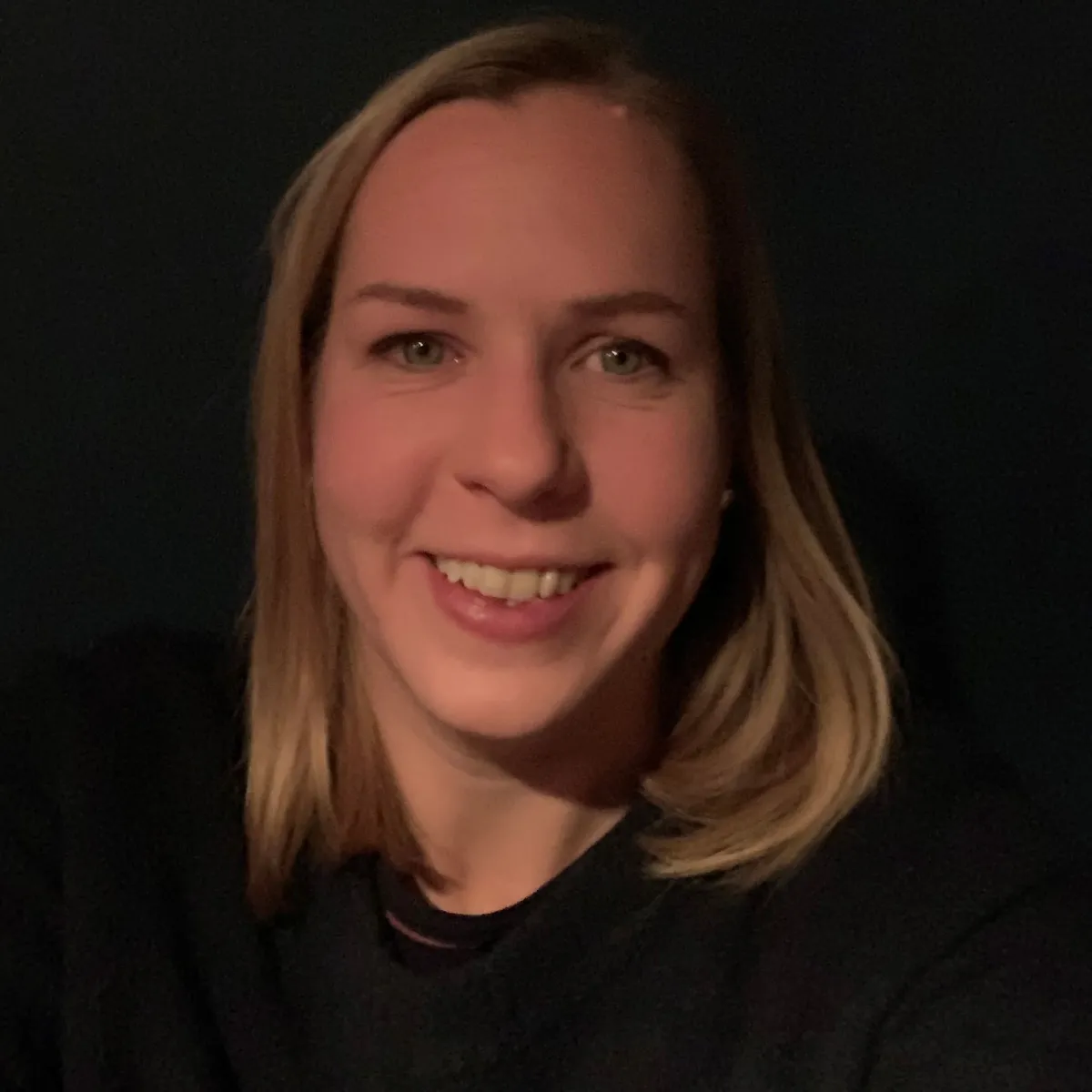 Headshot of adult female with light skin tone and blonde hair, with dark background