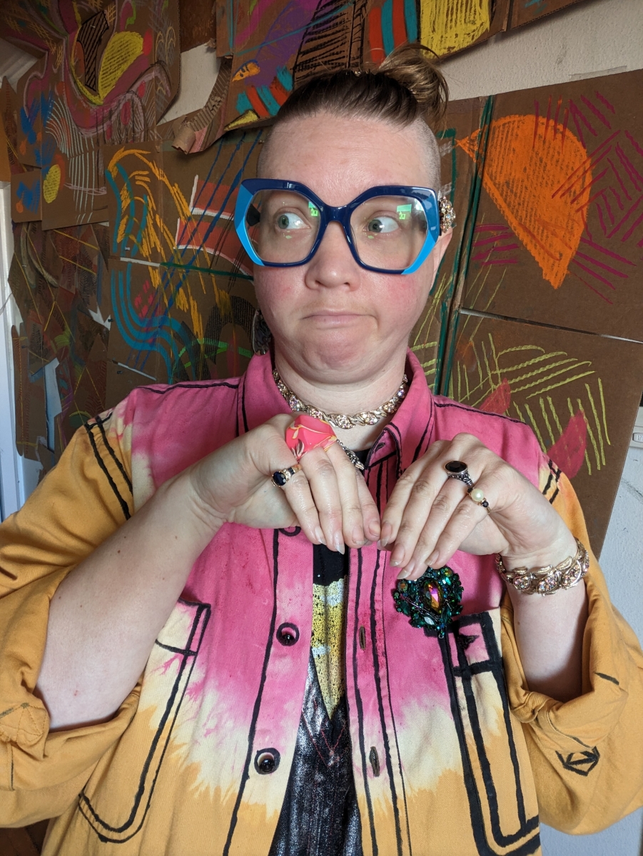A silly shot of a goofball artist wearing giant blue glasses and a pink and yellow button up in front of a colorful background.
