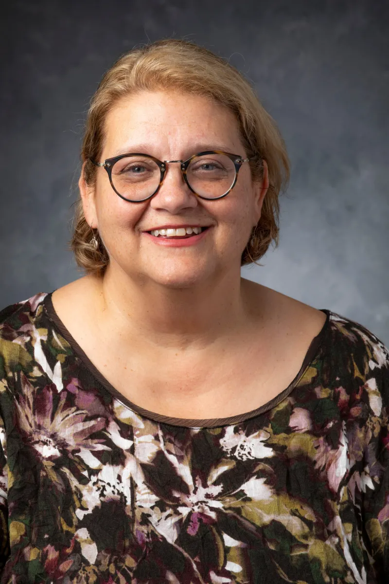 Headshot of a middle-aged white female with brown frame glasses, light brown hair to the neck, wearing a floral shirt with a black background.