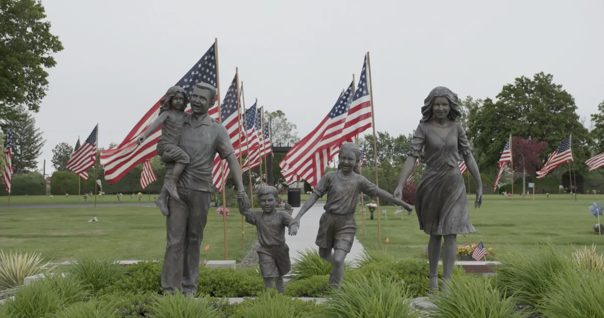 Still from 'Richland' depicting a statue of a family walking proudly in front of two columns of American flags, which border a series of manicured lawns. Photo credit: Helki Frantzen