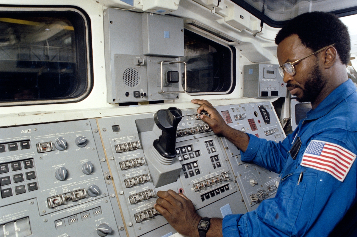 Still is from DC/DOX's Closing Night film 'The Space Race.' A Black man wearing glasses and a blue space suit with the American flag patch on the left arm, stands in front of a console on a space craft. 