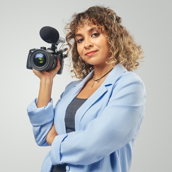 Headshot of a person with curly, shoulder length blonde hair holding a camera and smiling. They wear a blue blazer. 
