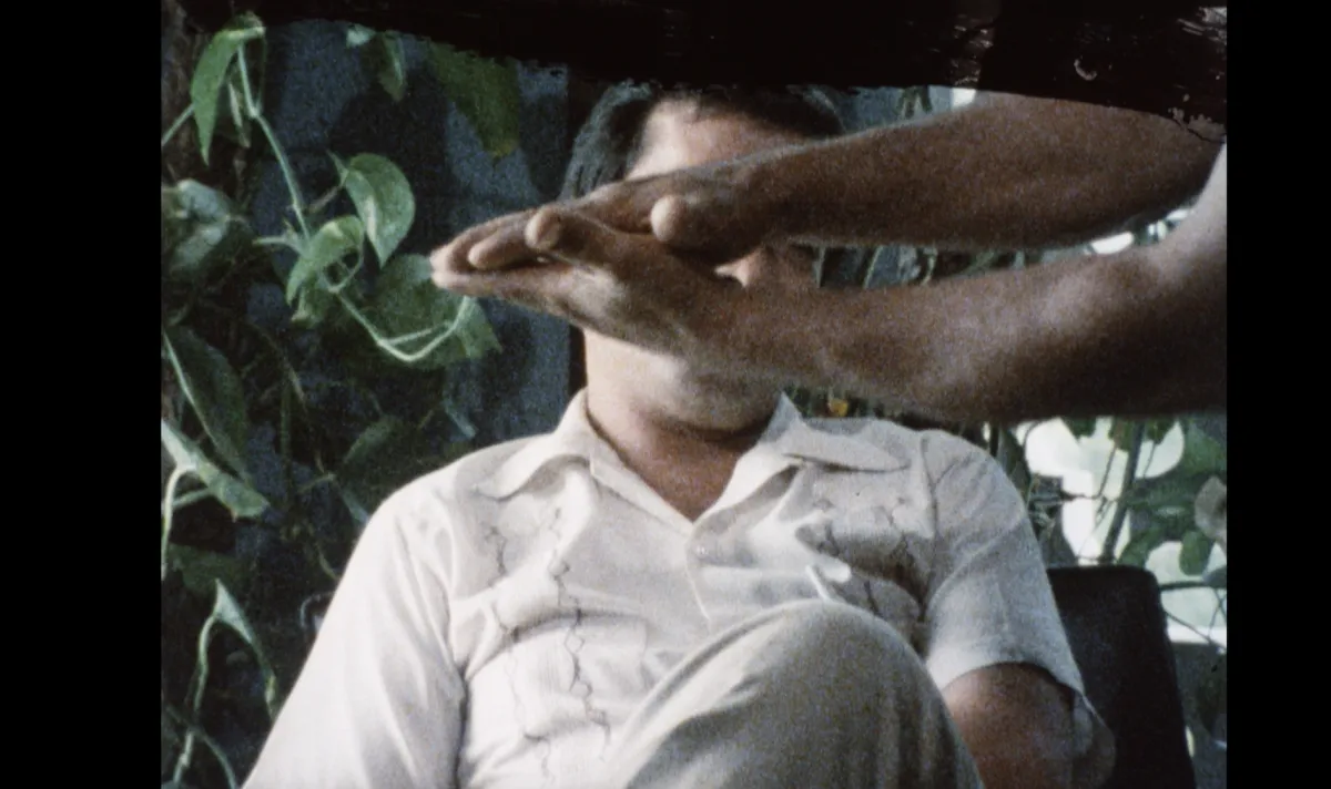 A man sits in a beige button up in a chair in front of greenery. In front of him hands and arms clasping, as if they are slating on a movie set.