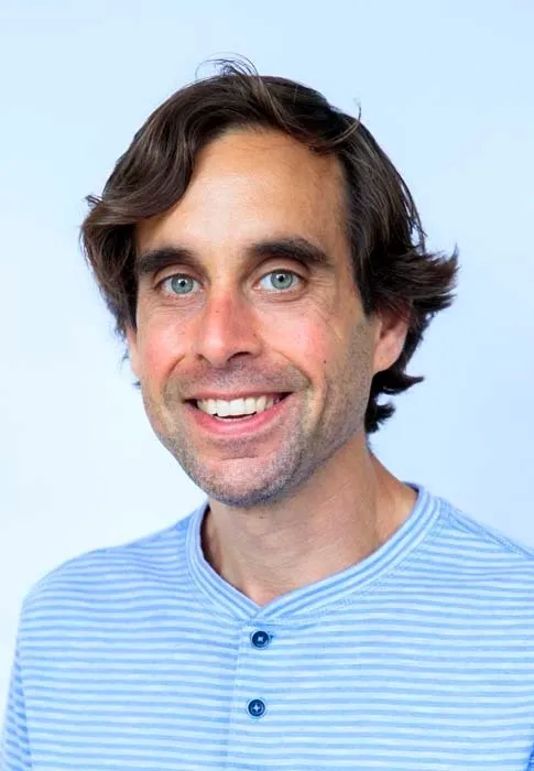 A man with dark brown hair, dark eyebrows and blue eyes smiles at the camera. He wears a shirt with blue strips and two buttons. 