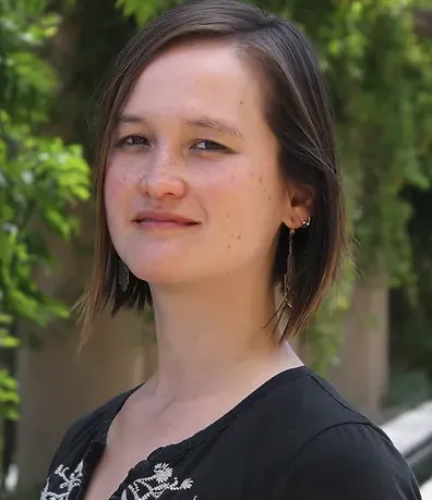 Headshot of a fair-skinned woman of mixed Asian and Caucasian descent with an angled bob, dangly gold earrings, and a black top, in front of leafy wall.