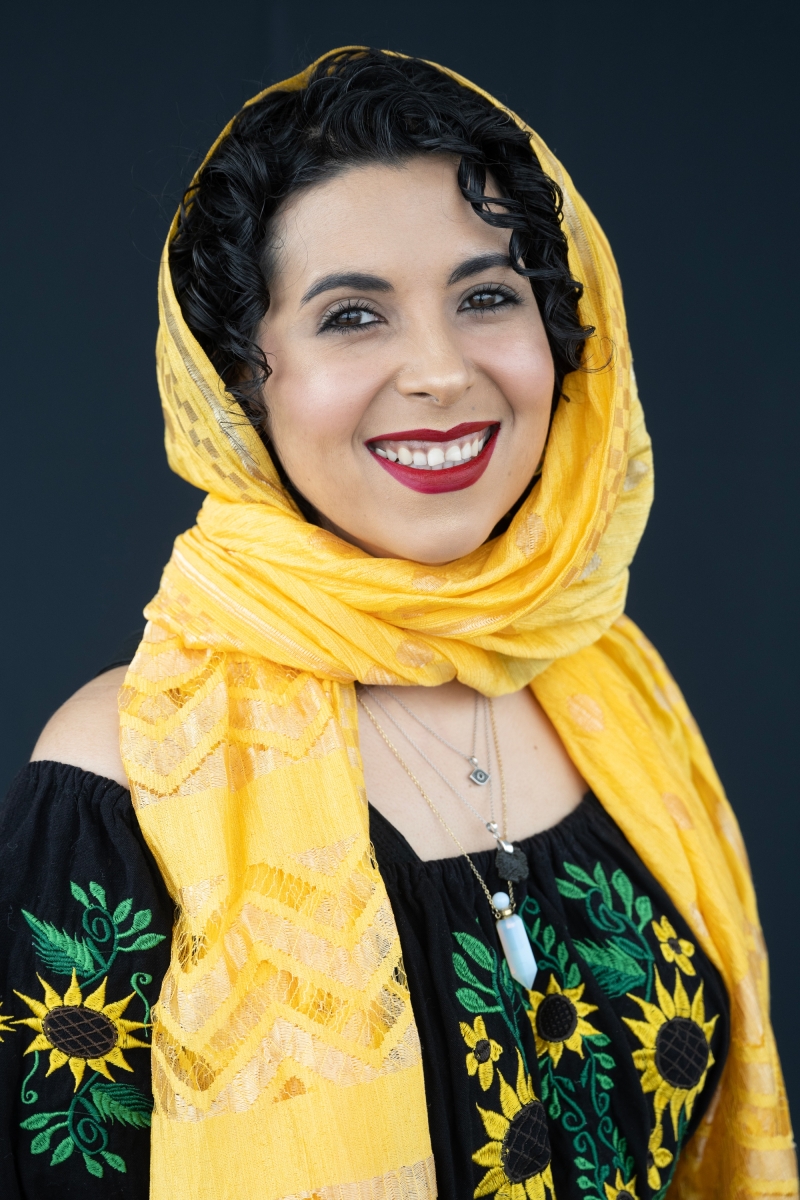 Headshot of an Indigenous and Middle Eastern Two-Spirit person with medium-light skin tone, curly black hair covered in a yellow head-scarf; They are wearing crimson lipstick and 3 layered semi-precious stone necklaces over a black shirt with sunflowers.