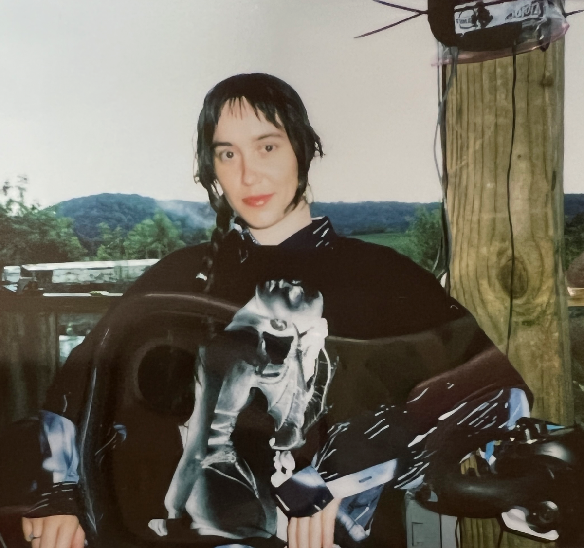 Poloroid of a white, non-binary person with dark brown hair and an Eartheater t-shirt, sitting in front of green hills.