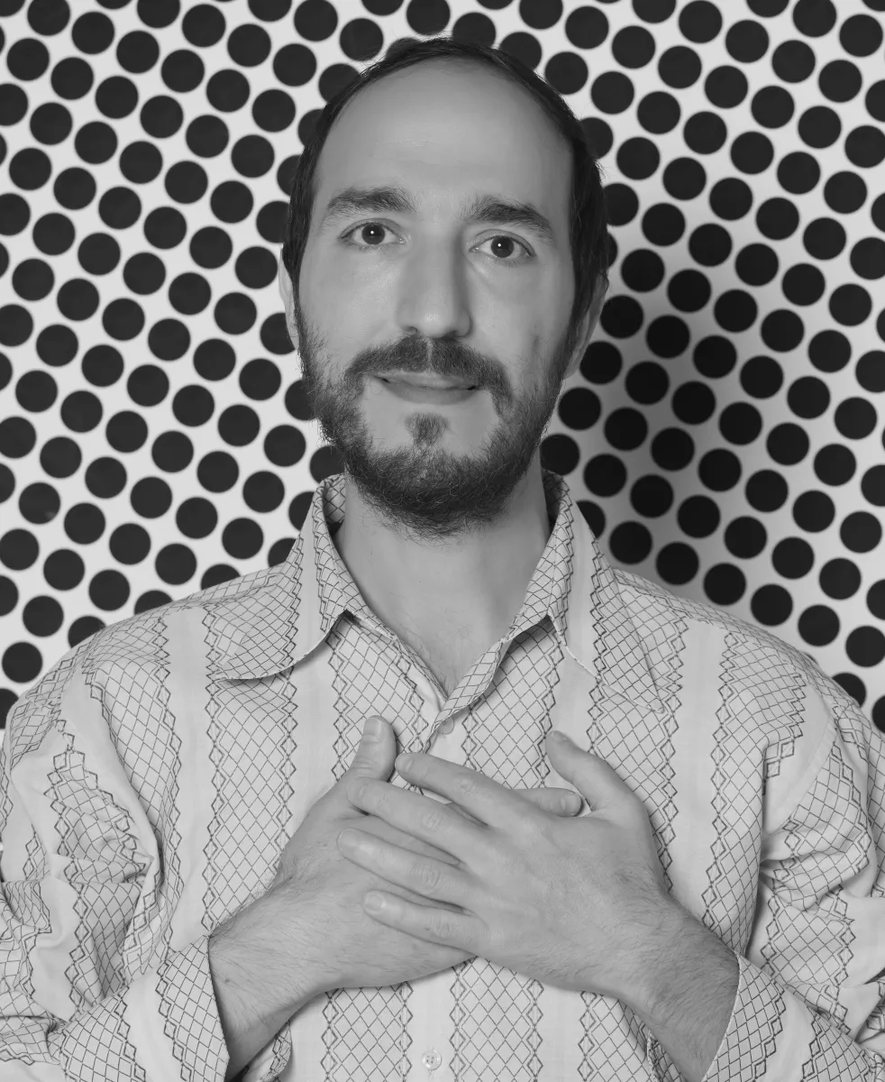 Black and white headshot of a middle-aged man with medium-light skin tone, trimmed beard and closed mouth smile, wearing a dress shirt with both hands on his chest.