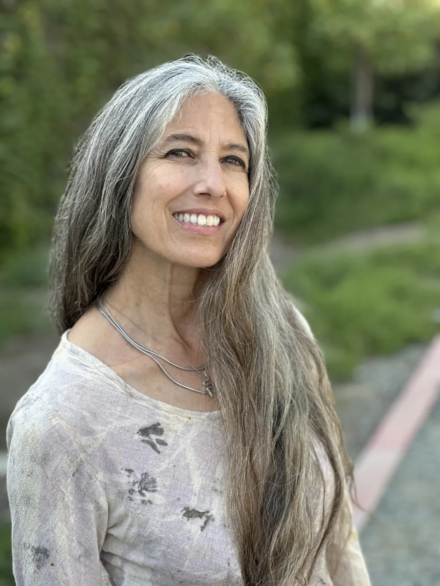 Headshot of a woman smiling with long gray and light brown hair wearing a mottled periwinkle with black splotches natural linen shirt in front of blurred green hedging plants