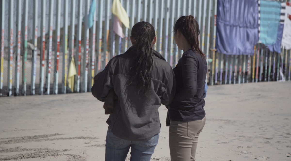 Photographed from behind, two people with long black hair stand in front of a tall, grey border wall.