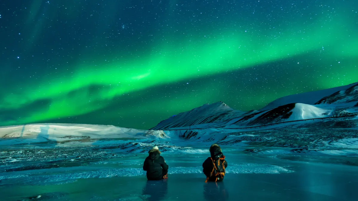 Two people, surely avid adventurers, stand waist deep in a body of arctic water; maybe they’re in Alaska - gazing upon a vast night sky. Most notably there is a bright, luminescent green aurora borealis shining above them. 