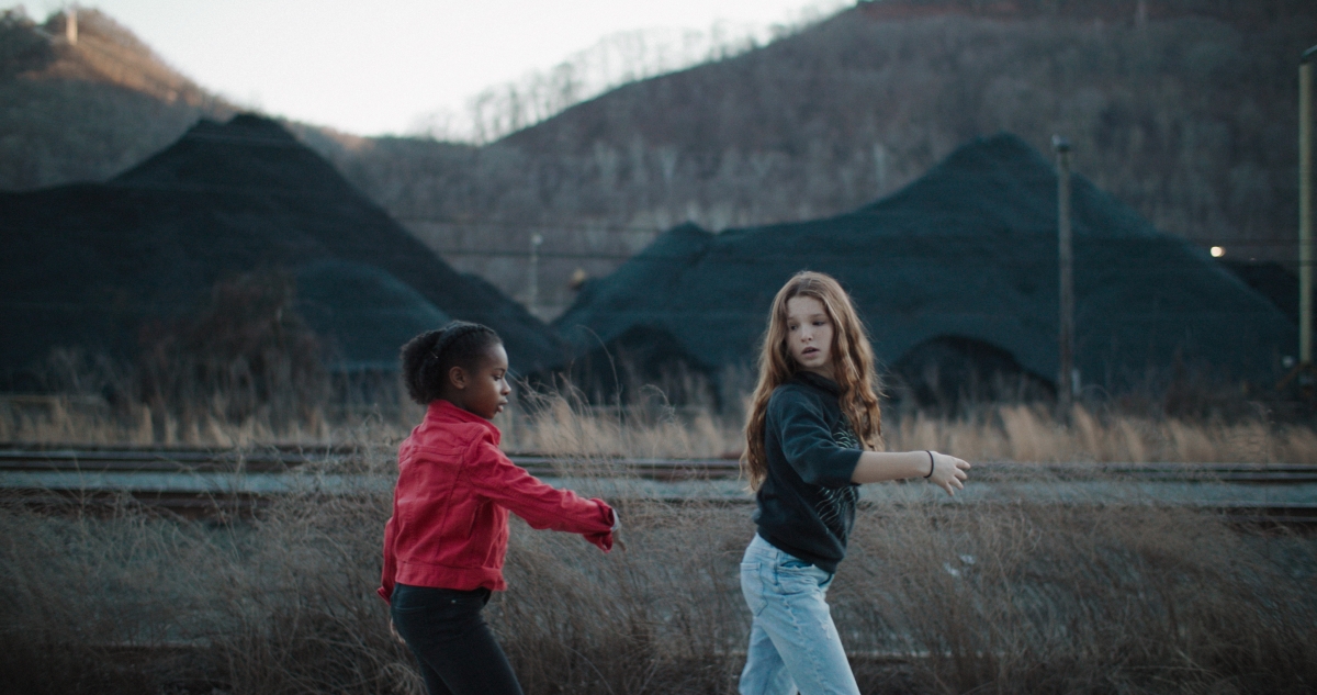Two young girls playing together against a backdrop of mountains, weeds, and mounds of dirt.