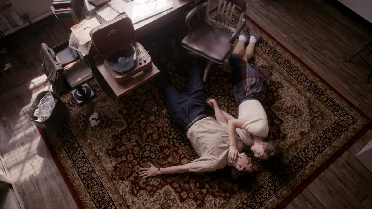 Two people lying on the floor in an office with dark brown hard wood floors, dark brown wooden chairs. One person is wearing long dark pants with a tan short sleeve button up shirt. The other person is wearing a dark plaid skirt with a white shirt sleeve top, ankles socks, with dark brown shoes.