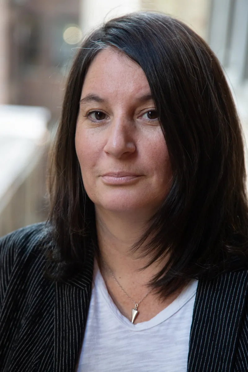 Headshot of an adult with long dark hair wearing a white T, black blazer and silver necklace.