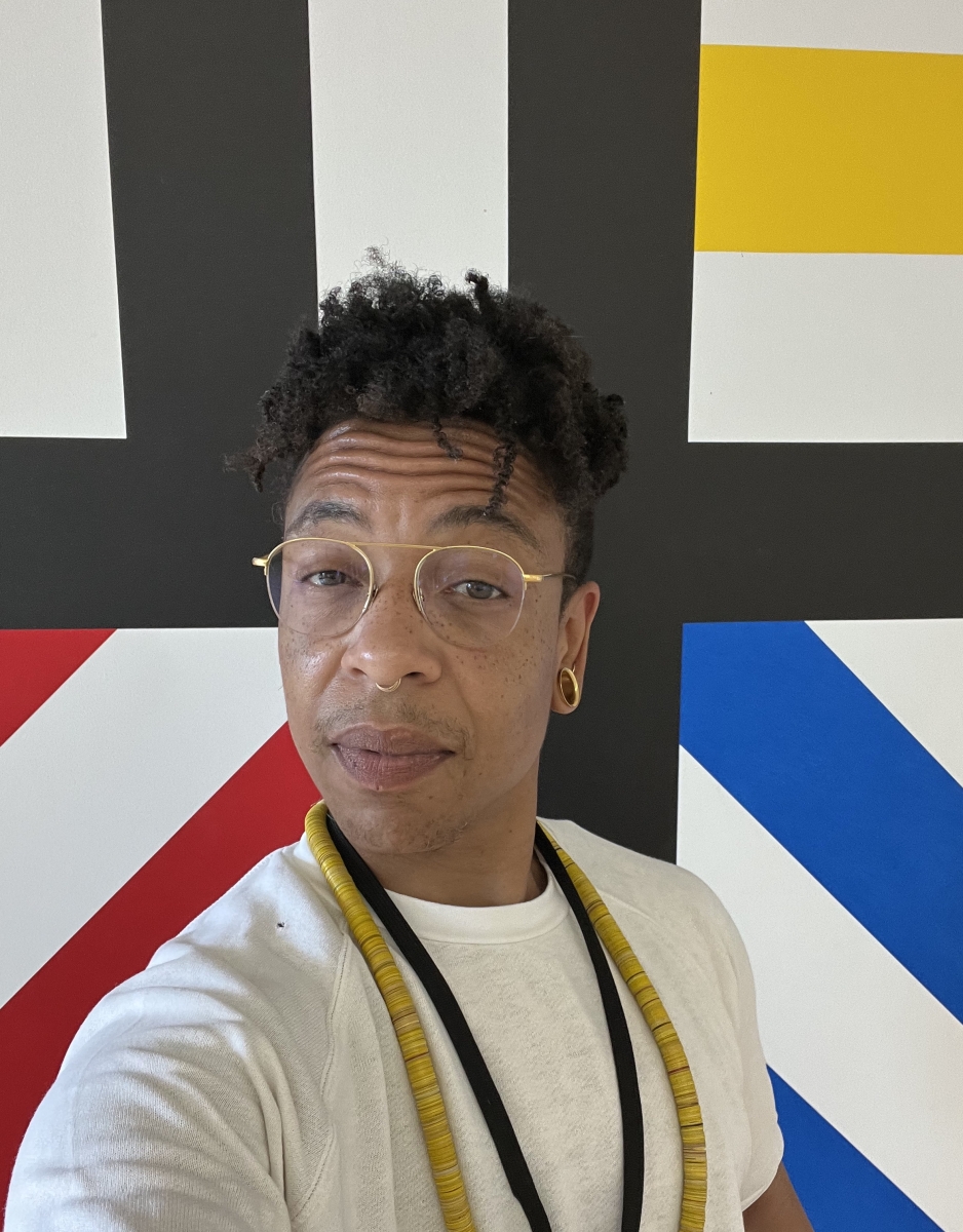 headshot of an adult male with a medium-light skin tone, wearing a white sweater, standing in front of a grid painting featuring bright red, yellow, blue, and black colors. 